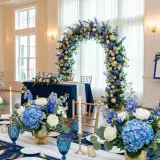 Elegancka sala weselna z niebieskim jako kolorem przewodnim. Kwiatowe łuki i dekoracje stołów w odcieniach błękitu i bieli.
