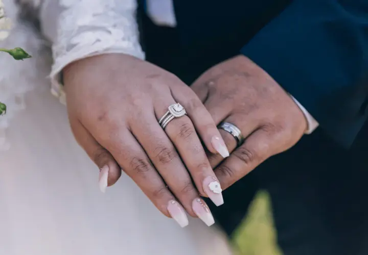 Dłonie nowożeńców z obrączkami i pięknymi paznokciami ślubnymi. Delikatny manicure zdobią kryształki i kwiat.