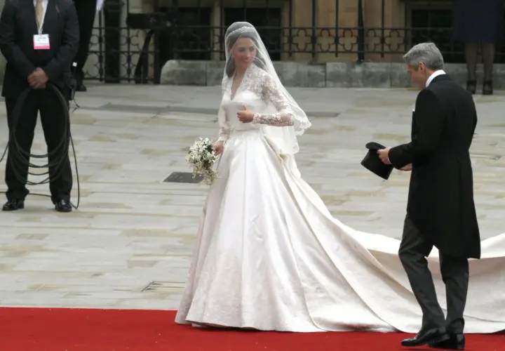 Kate Middleton w drugiej sukni ślubnej, z welonem i bukietem, idzie po czerwonym dywanie. Obok niej mężczyzna w żakiecie trzyma cylinder.