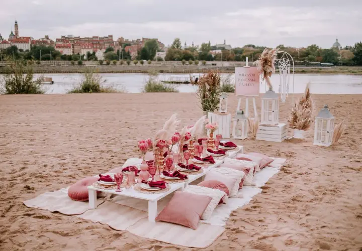 Elegancki wieczór panieński w plenerze nad rzeką. Stół z różowymi kieliszkami, serwetkami i kwiatami. W tle miasto.