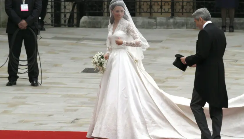 Kate Middleton w drugiej sukni ślubnej, z welonem i bukietem, idzie po czerwonym dywanie. Obok niej mężczyzna w żakiecie trzyma cylinder.
