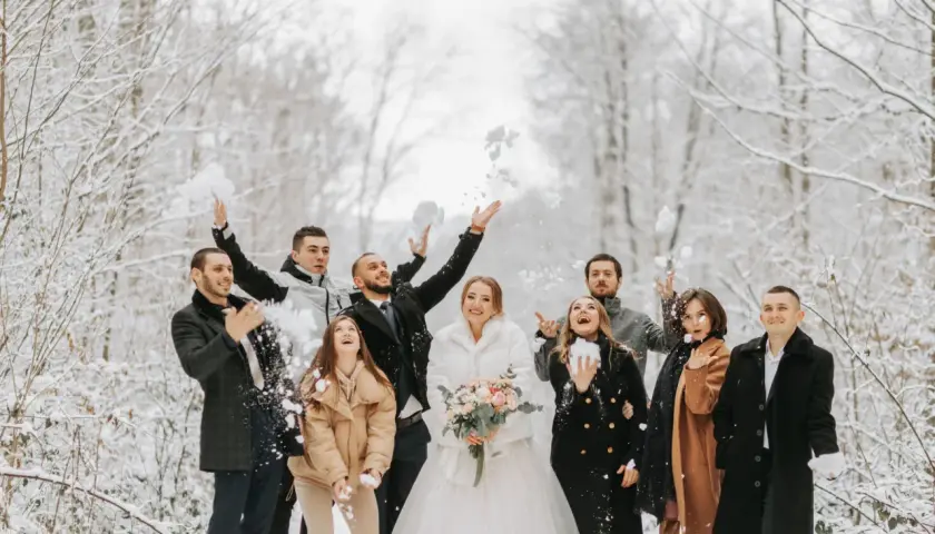 Para ślub zimą jak się ubrać? Para młoda i goście w zimowym lesie rzucają śnieżkami. Panna młoda w białym futerku, pan młody w eleganckim płaszczu.