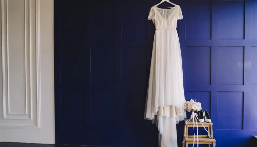 Elegancka suknia ślubna wisi na wieszaku, obok bukiet i buty. To inspiracja, jak przechowywać suknię ślubną, by zachowała swój blask.