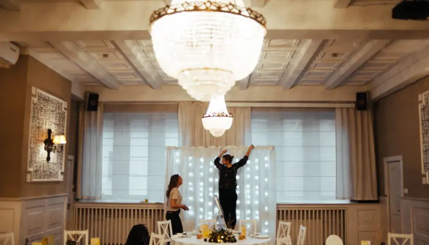 Dwie kobiety przygotowują salę weselną, jedna stoi na drabinie, dekorując tło światełkami. To początek drogi, jak zostać wedding plannerką.