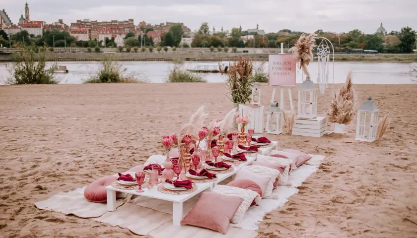Elegancki wieczór panieński w plenerze nad rzeką. Stół z różowymi kieliszkami, serwetkami i kwiatami. W tle miasto.