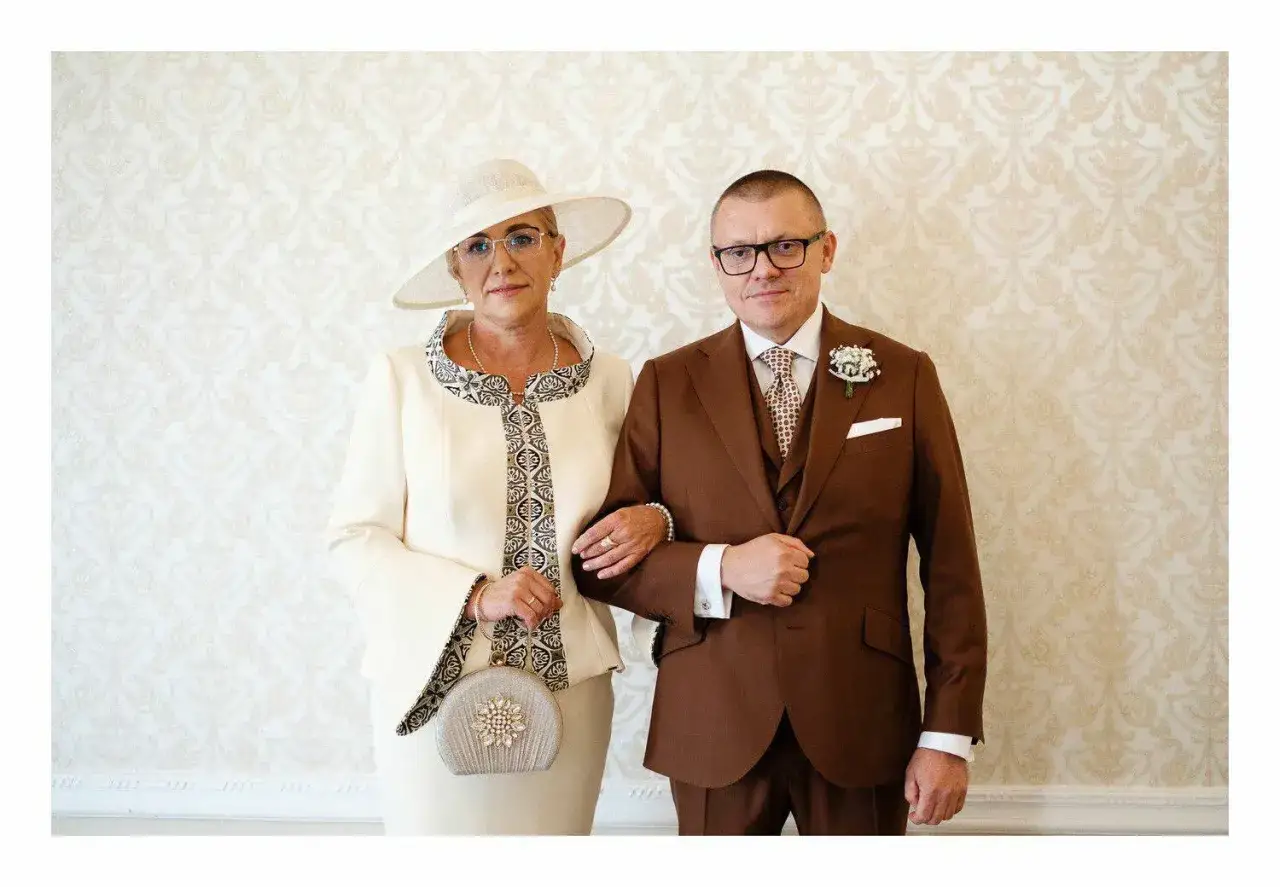 Elegancka matka panny młodej w kremowym stroju i kapeluszu, obok mężczyzny w brązowym garniturze.